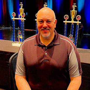 Dave Schreier smiling at the camera in a red shirt, gold and blue trophies in the background